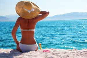 A woman in a white bikini and wide-brimmed hat sits on the beach facing the ocean