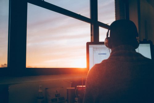 A person works on a computer while wearing headphones and sitting in front of a large window at sunset.