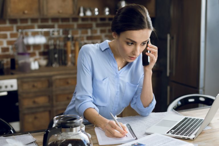 A woman in a blue shirt speaks on a phone while writing on a piece of paper